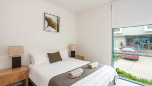 A hotel room with a large bed, two bedside tables, a lamp on each, a painting on the wall, and a window with a view of a red car and a building outside.