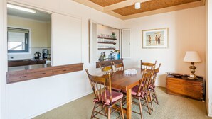 A dining area with a wooden table and chairs, a mirror, a shelf with glasses and bottles, and a painting on the wall.