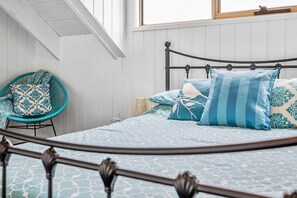 A bed with a metal frame and patterned bedding, a wicker chair with cushions, and a window above the bed.