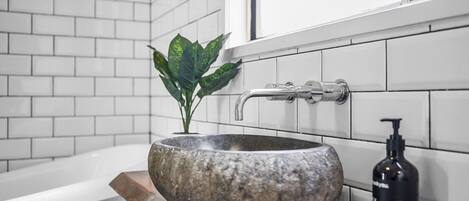 Um banheiro com pia de pedra, uma torneira, uma embalagem de sabonete e um vaso com planta.