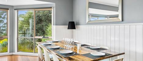 Un coin repas avec une table en bois dressée pour six personnes, des chaises blanches et une grande fenêtre donnant sur un paysage verdoyant.