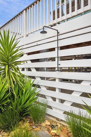 A white wooden fence with a built-in showerhead, surrounded by green plants and rocks.