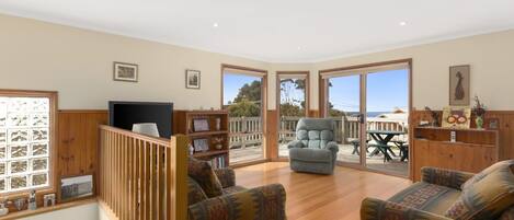 A spacious living area with a wooden floor, two patterned sofas, a television, and a balcony with a view of the sea.