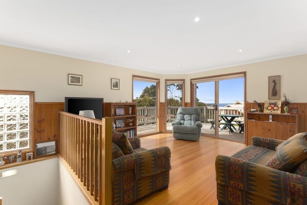 A spacious living area with a wooden floor, two patterned sofas, a television, and a balcony with a view of the sea.