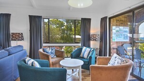 Una sala de estar moderna con un sofá azul, sillones verde mar y una mesa de centro blanca. Amplias ventanas ofrecen vistas a árboles y un edificio de ladrillo.