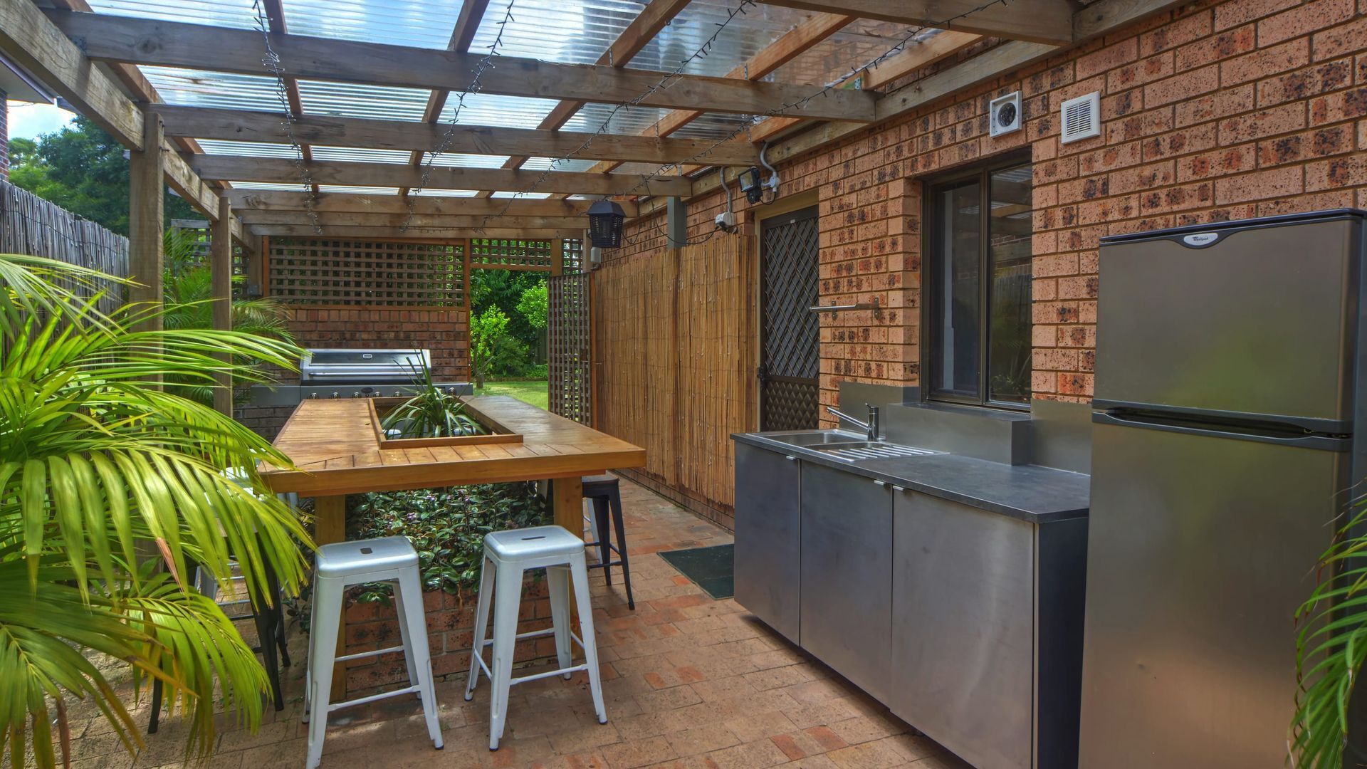 A covered outdoor kitchen with a grill, bar stools, and a refrigerator.
