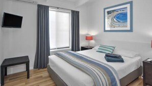 Una habitación de hotel con cama, mesita de noche, televisor montado en la pared y ventana con cortinas.