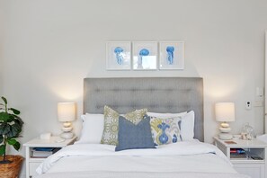 A neatly made bed with a grey upholstered headboard, flanked by two bedside tables with lamps and a potted plant.