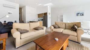Ein modernes Wohnzimmer mit zwei beigefarbenen Sofas, einem Holztisch und einer Küche im Hintergrund.