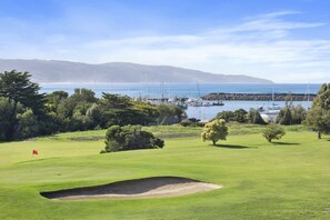 A golf course with a bunker, trees, and a view of a marina with boats and a distant mountain range. - Apollo Blue 11 (Apollo Bay)