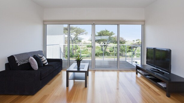 A modern living room with a dark sofa, a small table with a plant, a flat-screen TV, and a sliding glass door leading to a balcony with a view of greenery. - Four Kings 3 (Anglesea)