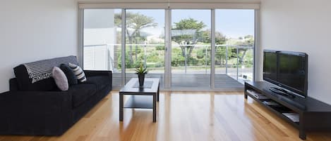 A modern living room with a dark sofa, a small table with a plant, a flat-screen TV, and a sliding glass door leading to a balcony with a view of greenery.