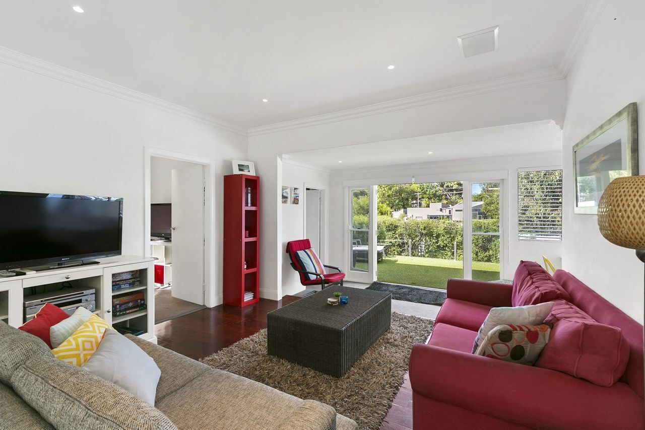 Una sala moderna con televisor de pantalla plana, sofás rojo y gris, una estantería roja y un amplio ventanal con vistas al exterior.
