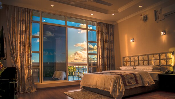 A bedroom with a large window, a bed, a nightstand, and a view of the outdoors. - Abuharee Grand (Hithadhoo)