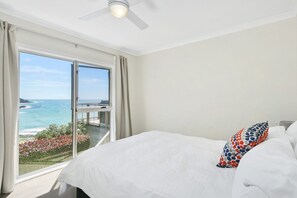A bedroom with a large window, a bed with white bedding, and a view of the sea.