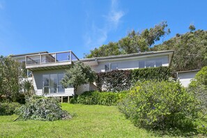 A modern two-story house with a balcony, surrounded by lush greenery and trees.