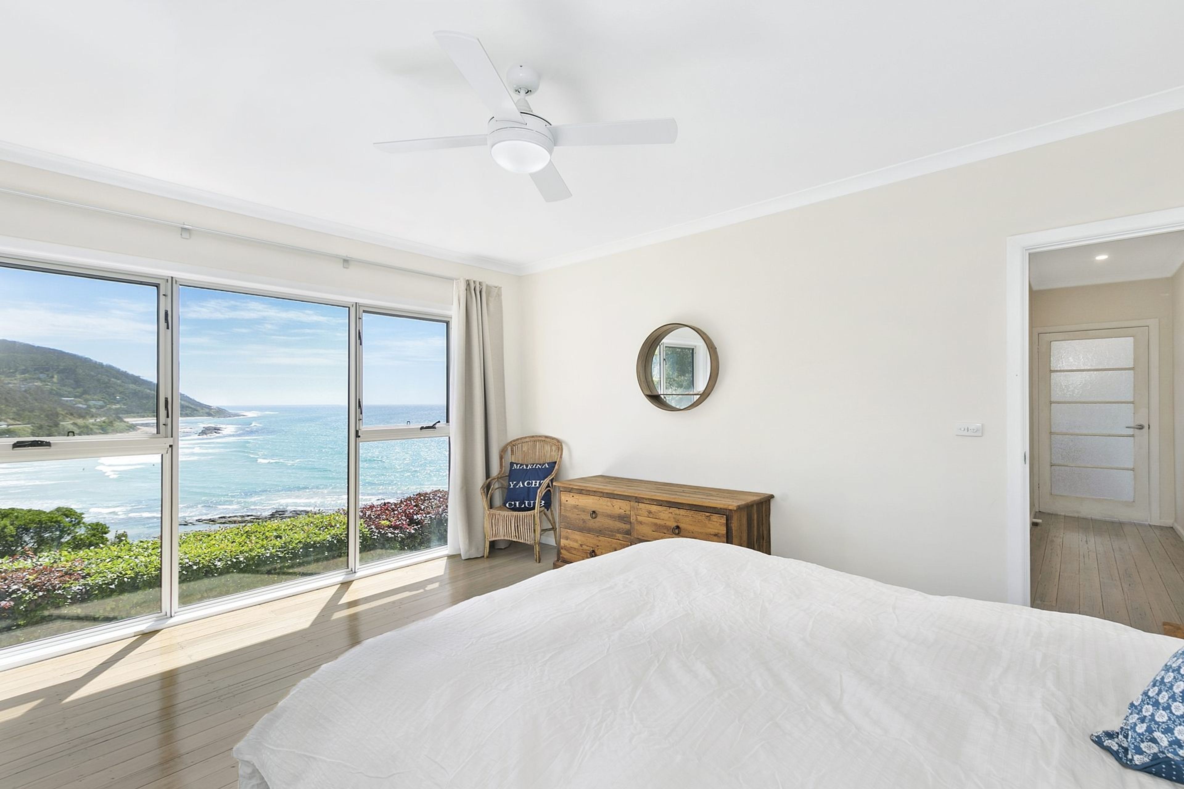 Une chambre avec une grande fenêtre, un lit, une commode en bois, une chaise et un ventilateur de plafond.