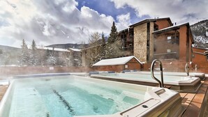 Zona de hidromasaje con una terraza de madera, un edificio de piedra al fondo y árboles cubiertos de nieve.