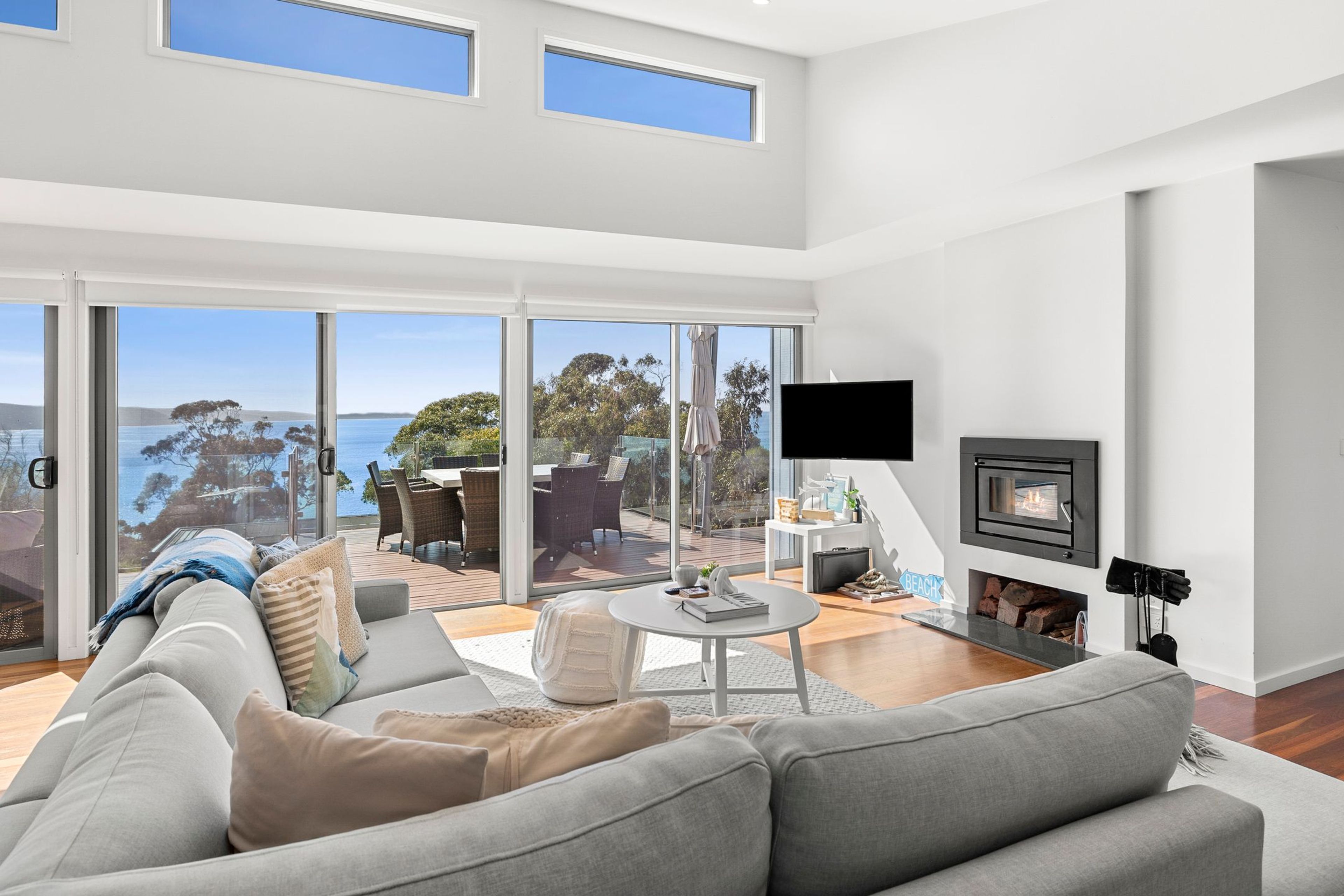 A modern living room with a large sofa, a coffee table, and a flat-screen TV. There is a fireplace and a view of the ocean through large windows.