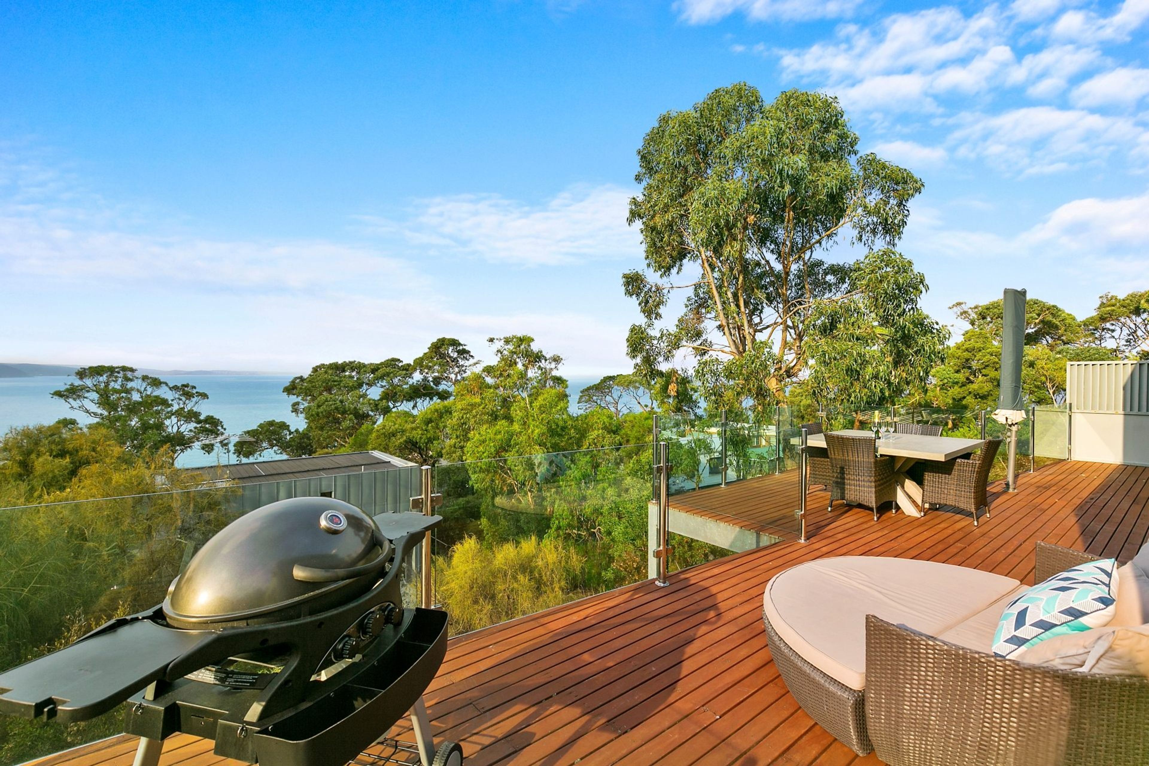 A deck with a barbecue grill, outdoor furniture, and a view of the sea and trees.