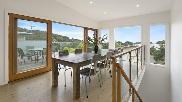 A modern dining area with a wooden table and chairs, large windows with a view of greenery, and a balcony with outdoor seating. - Godalming (Lorne)