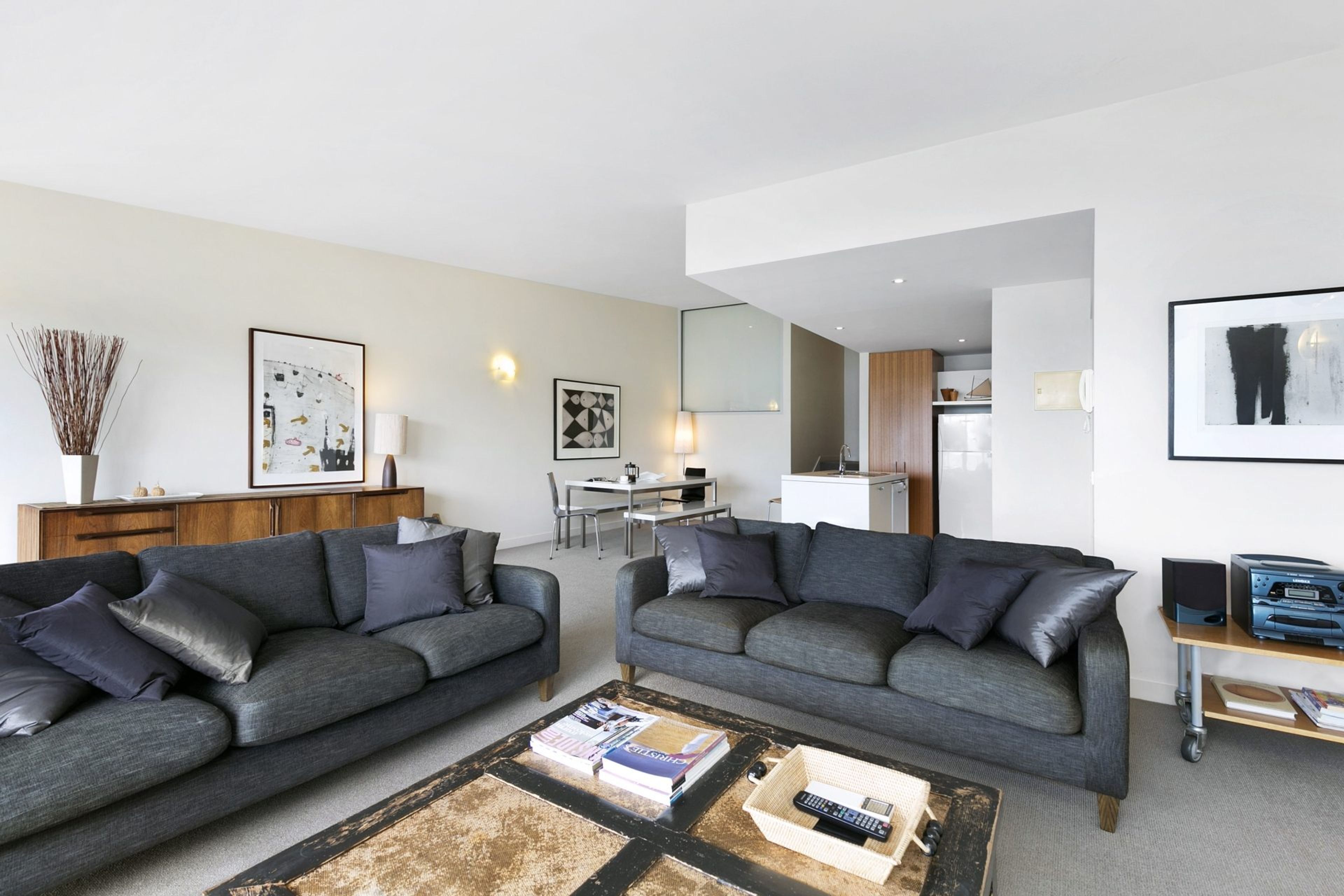 Un salon moderne avec deux canapés gris, une table basse avec des livres et des télécommandes, un coin repas avec une table et des chaises, et une cuisine en arrière-plan.
