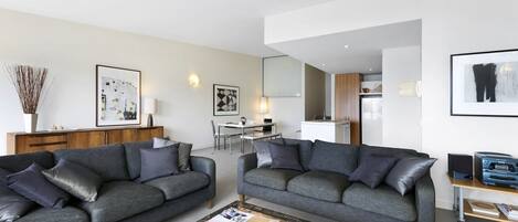 Un salon moderne avec deux canapés gris, une table basse avec des livres et des télécommandes, un coin repas avec une table et des chaises, et une cuisine en arrière-plan.