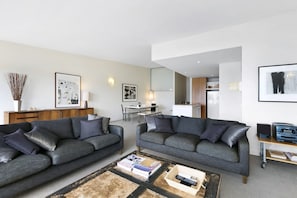 A modern living room with two grey sofas, a coffee table with books and remote controls, a dining area with a table and chairs, and a kitchen in the background.
