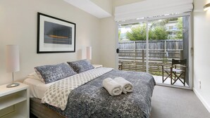 A bedroom with a bed, a nightstand, a lamp, a framed picture on the wall, and a view of the outside through a sliding glass door. - Apartment 25 Kalimna (Lorne)