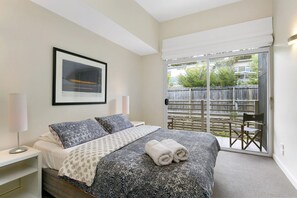 A bedroom with a bed, a nightstand, a lamp, a framed picture on the wall, and a view of the outside through a sliding glass door. - Apartment 25 Kalimna (Lorne)