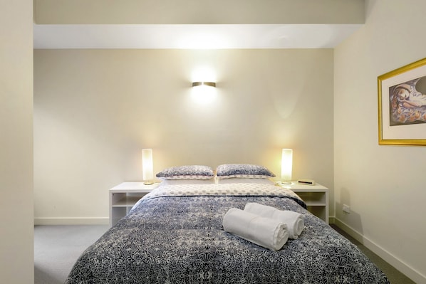 A single bed with patterned bedding, two bedside lamps, a bedside table, and a framed picture on the wall.