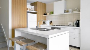A modern kitchen with a central island, white cabinetry, and a wooden accent wall. - Apartment 25 Kalimna (Lorne)