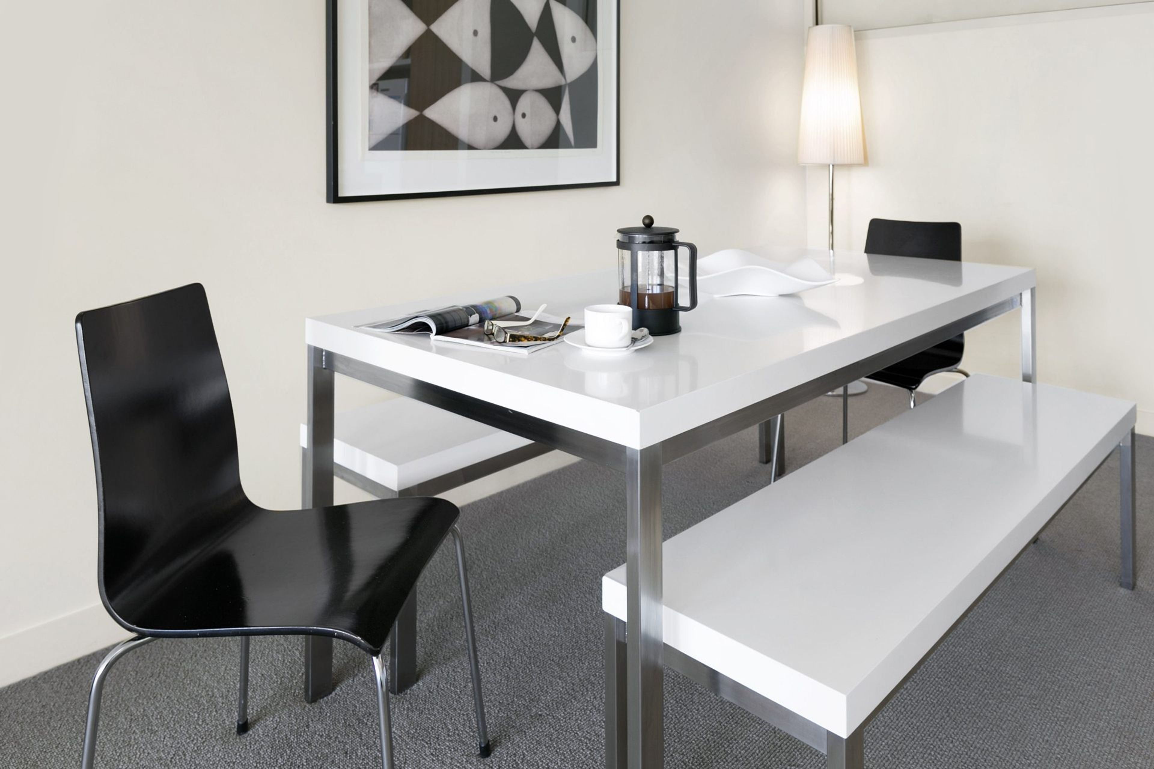 Een moderne hotelkamer met een witte tafel, zwarte stoelen, een koffiezetapparaat, een ingelijste foto en een lamp.