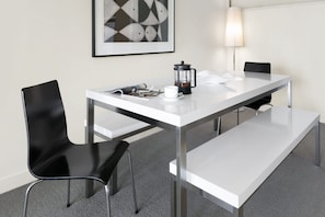 A modern hotel room with a white table, black chairs, a coffee maker, a framed picture, and a lamp.