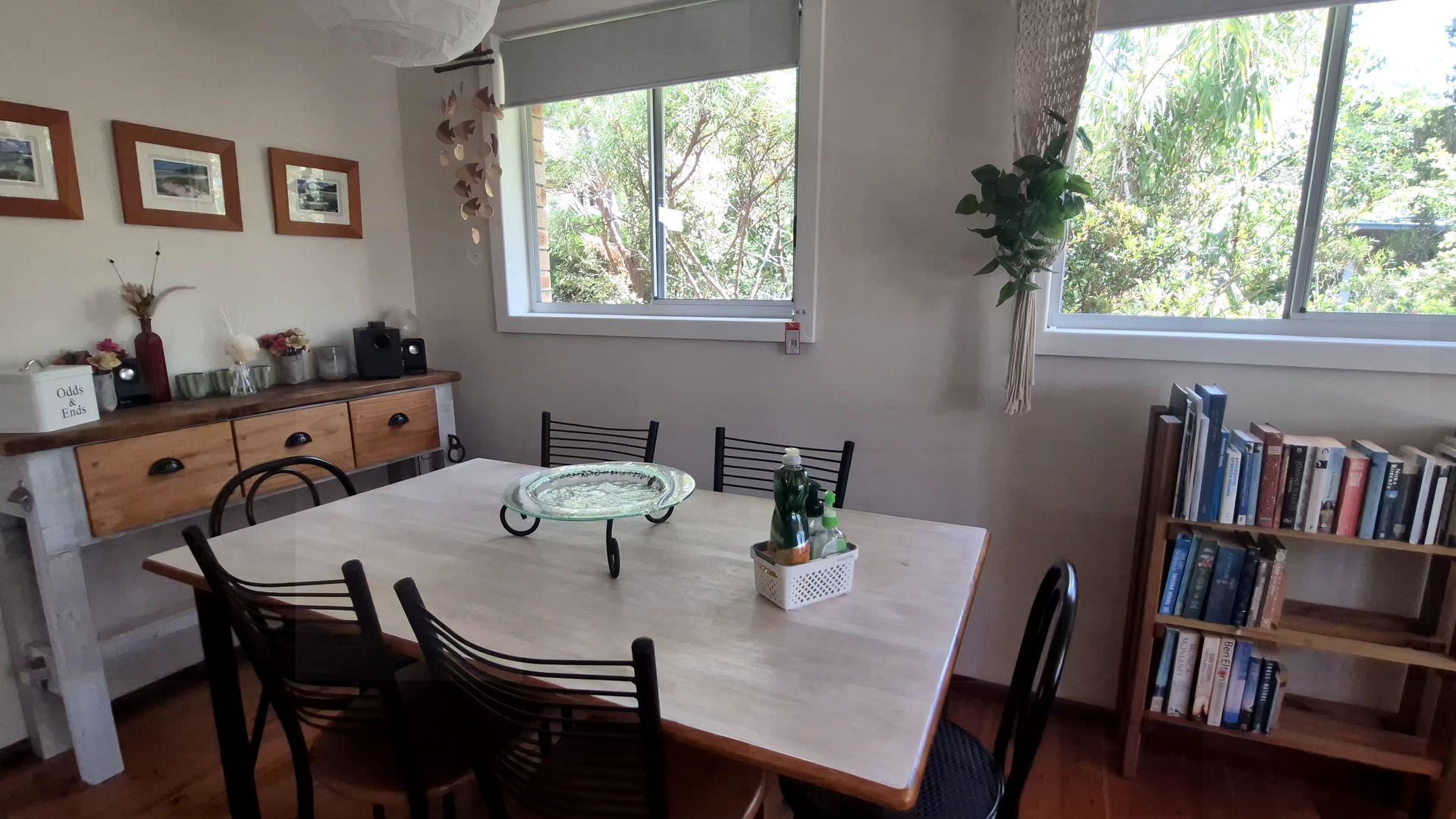 A dining area with a wooden table, chairs, a glass-top table centerpiece, a bookshelf, and a window with a view of trees.