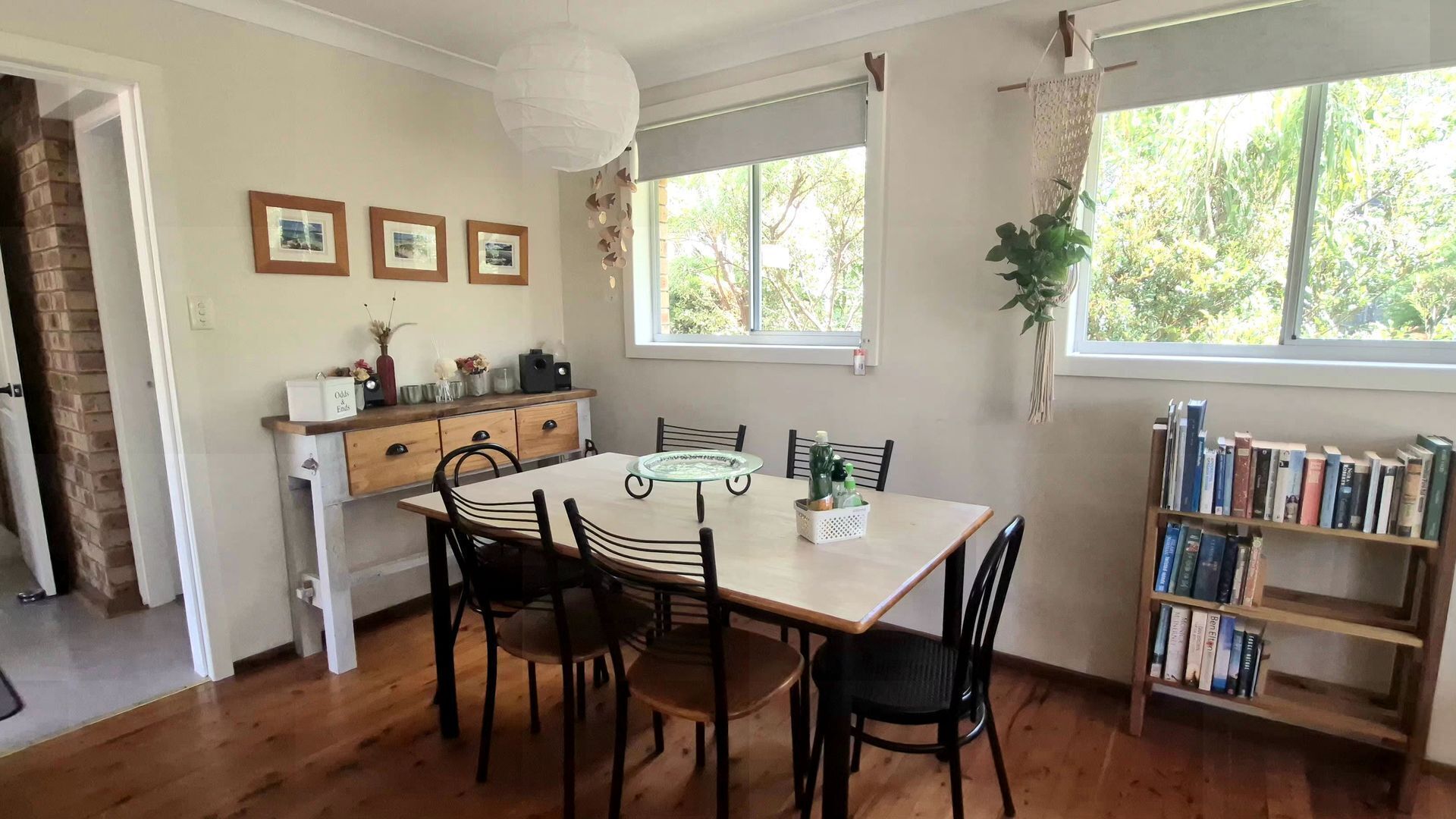 A dining area with a wooden table and chairs, a bookshelf with books, and a window with a view of trees.