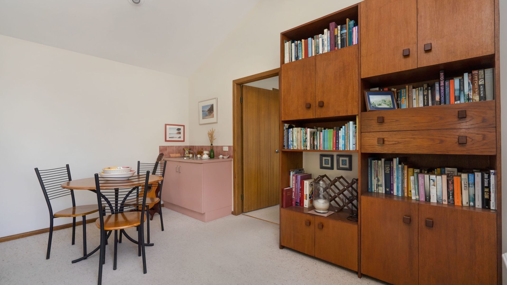 A room with a dining table, chairs, a kitchen area, and a large wooden bookshelf filled with books.