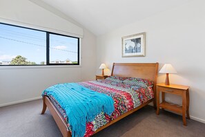 A bedroom with a large window, a bed with a colorful blanket, two bedside tables with lamps, and a framed picture on the wall.