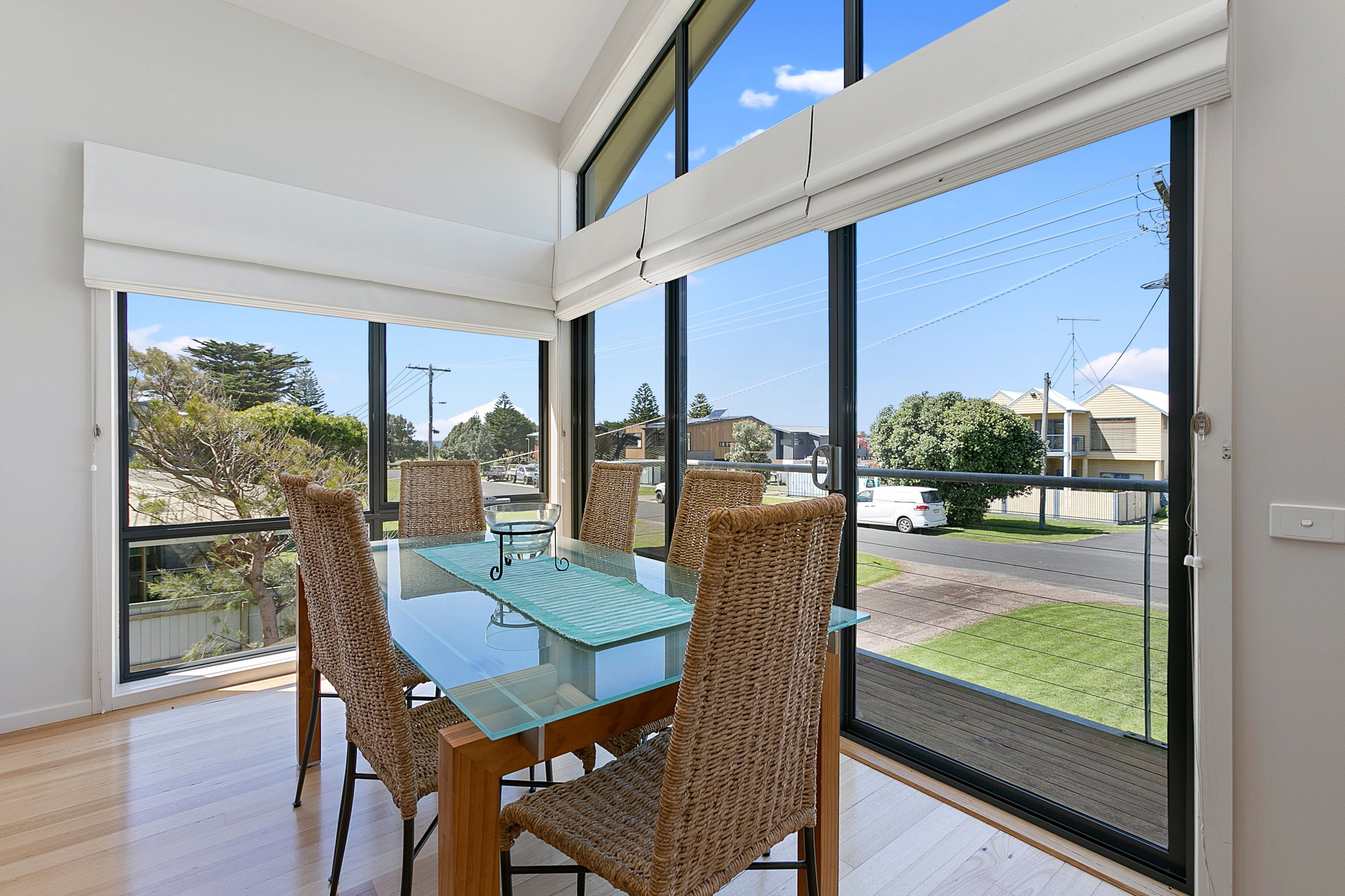 A dining area with a glass table and wicker chairs, large windows offering a view of the street and greenery.
