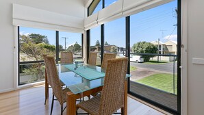 Un coin repas avec une table en verre et des chaises en osier, de grandes fenêtres donnant sur la rue et la verdure.