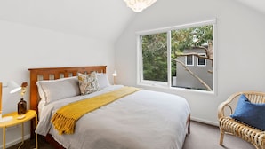 A bedroom with a bed, a bedside table, a lamp, a window with a view of trees, and a wicker chair.