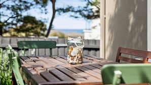 A wooden table with a glass jar containing seashells and a teddy bear, set outdoors with a wooden fence and greenery in the background.
