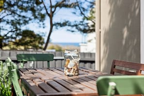 A wooden table with a glass jar containing seashells and a teddy bear, set outdoors with a wooden fence and greenery in the background.