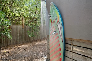 A surfboard leaning against a wall in a shaded area with a wooden fence and mulch ground.
