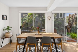 A dining area with a wooden table, grey upholstered chairs, and a view of a garden through large sliding glass doors.