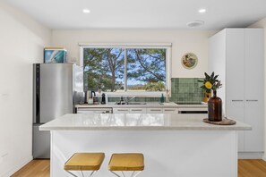A modern kitchen with a central island, two stools, a refrigerator, and a window with a view of trees and a building.
