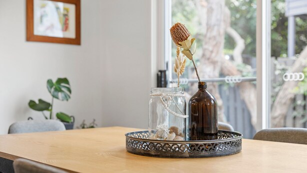 Et bord med et dekorativt glas og en mørk flaske, et billede i ramme på væggen og en plante i baggrunden.