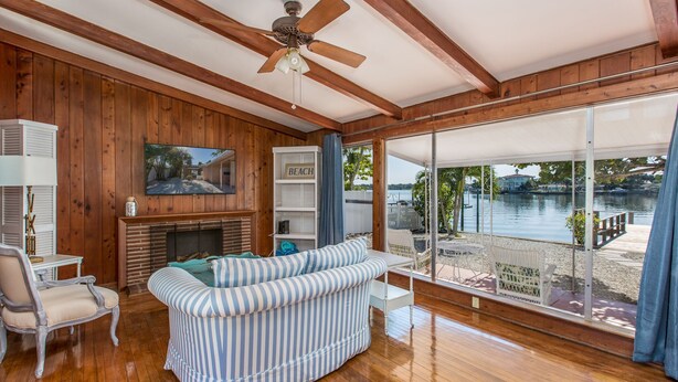 A spacious living room with wooden walls, a fireplace, a large window overlooking a lake, and a ceiling fan. - 🌴 Charming 2BR WATERFRONT Cottage - Perfect for a Coastal Getaway! (Indian Rocks Beach)