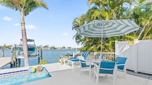A pool area with a table and chairs, a striped umbrella, and a view of the water. - Water Lover's Paradise | Waterfront • Private Dock • Heated Pool (Indian Rocks Beach)