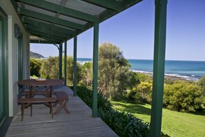 A covered outdoor area with a wooden table and benches, overlooking a coastal landscape with vegetation and the sea.